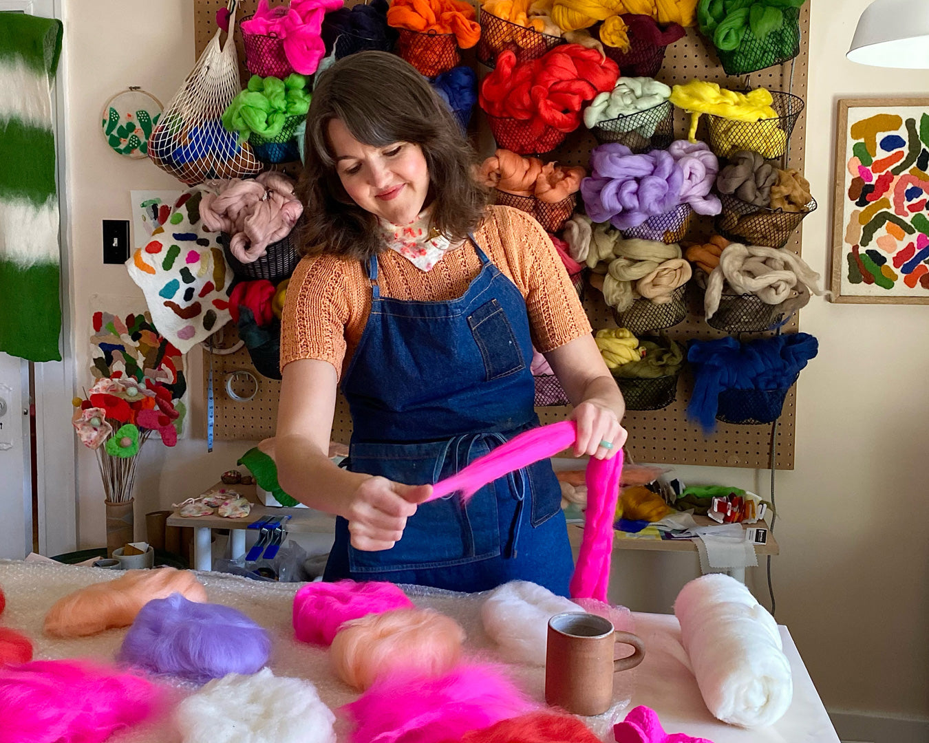 Artisan stretching vibrant pink wool during water felting workshop, surrounded by a rainbow of wool supplies.