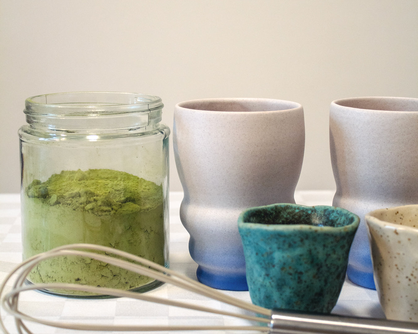 A jar of matcha powder next to ceramic cups and a whisk, ready for a matcha latte recipe.