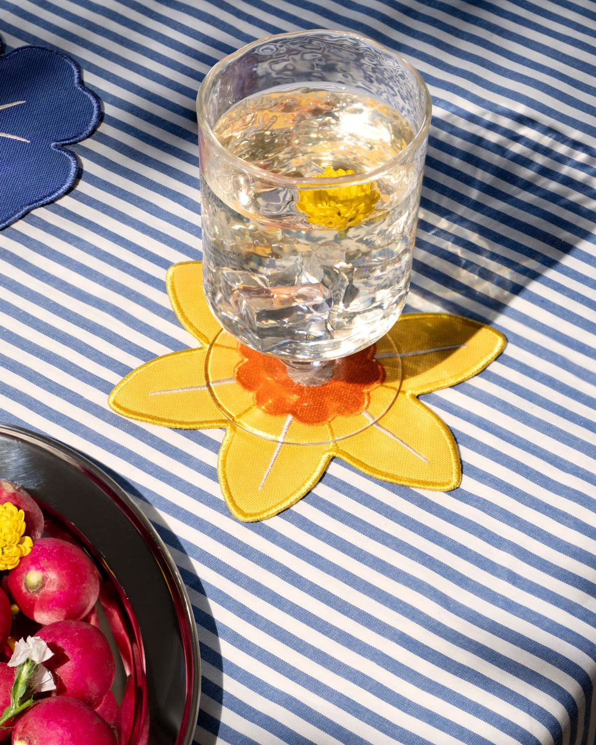 A glass of water with a yellow flower rests on Octaevo Daffodil Coasters (Set of 4) atop a blue striped tablecloth.