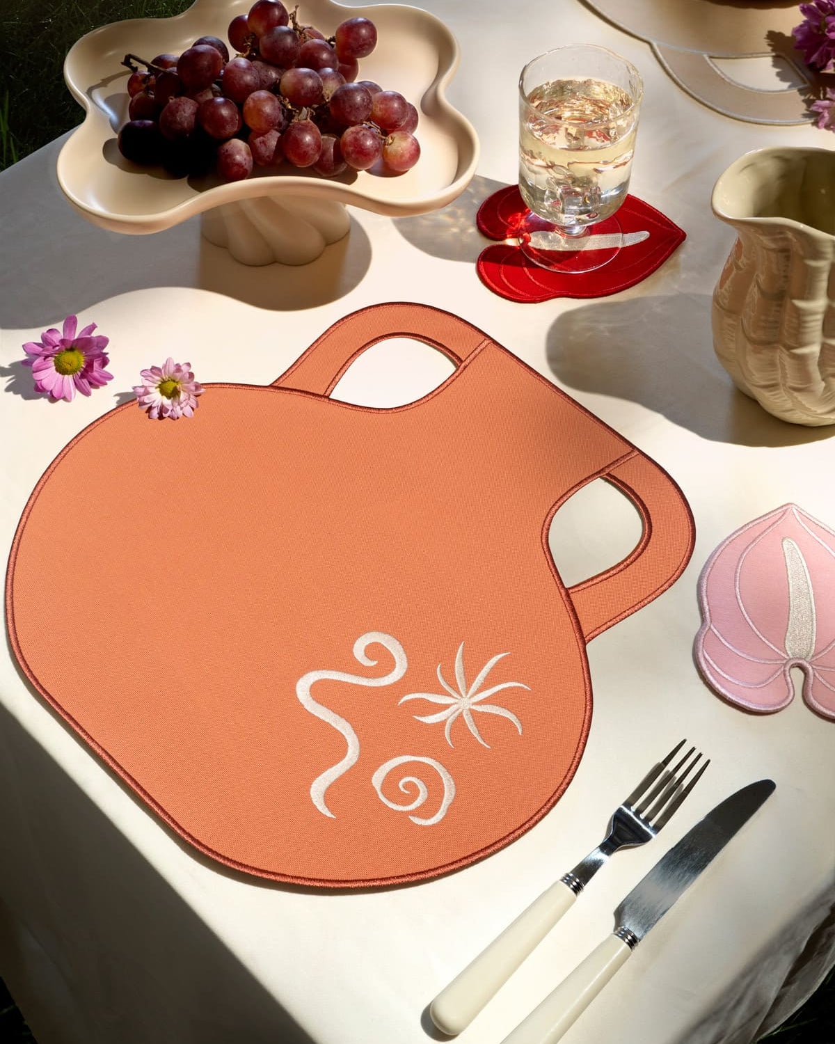 An outdoor table with grapes, a drink, flowers, and Octaevo’s Terracotta Amphora Placemats (Set of 2).