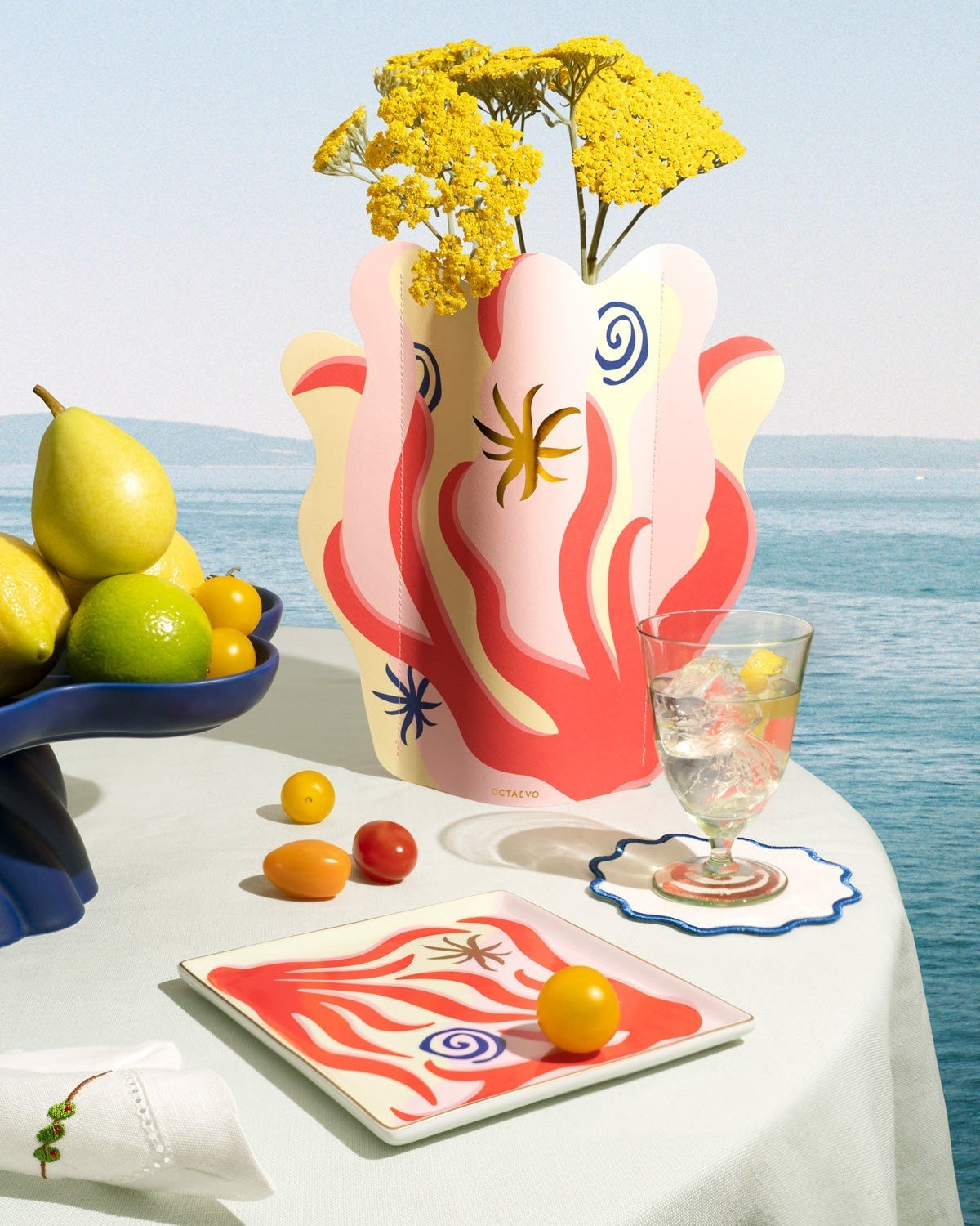 A colorful table setting with fruit, a floral vase, a glass of water, and a plate, set against a seaside background.