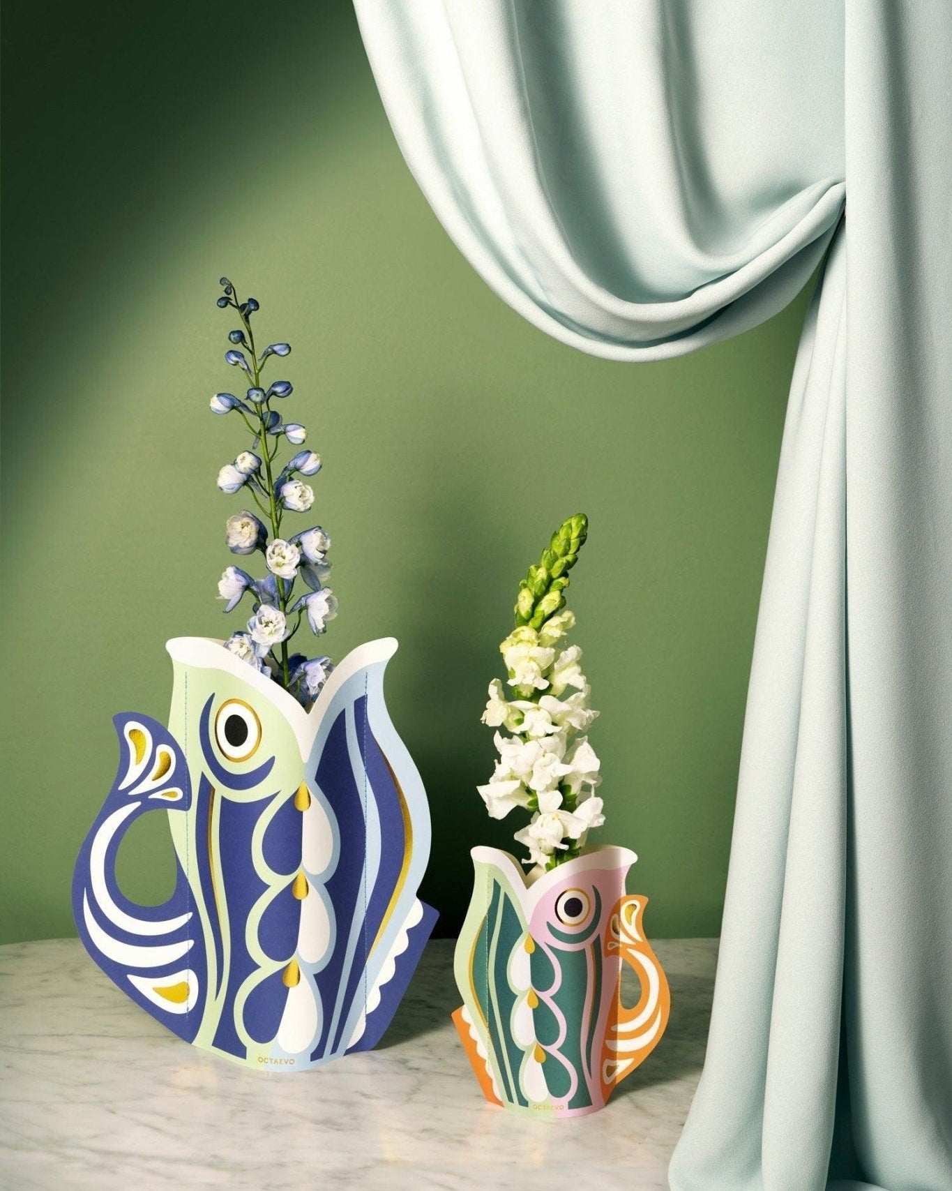 Two decorative OCTAEVO paper vases in orange with floral arrangements against a green curtain background.