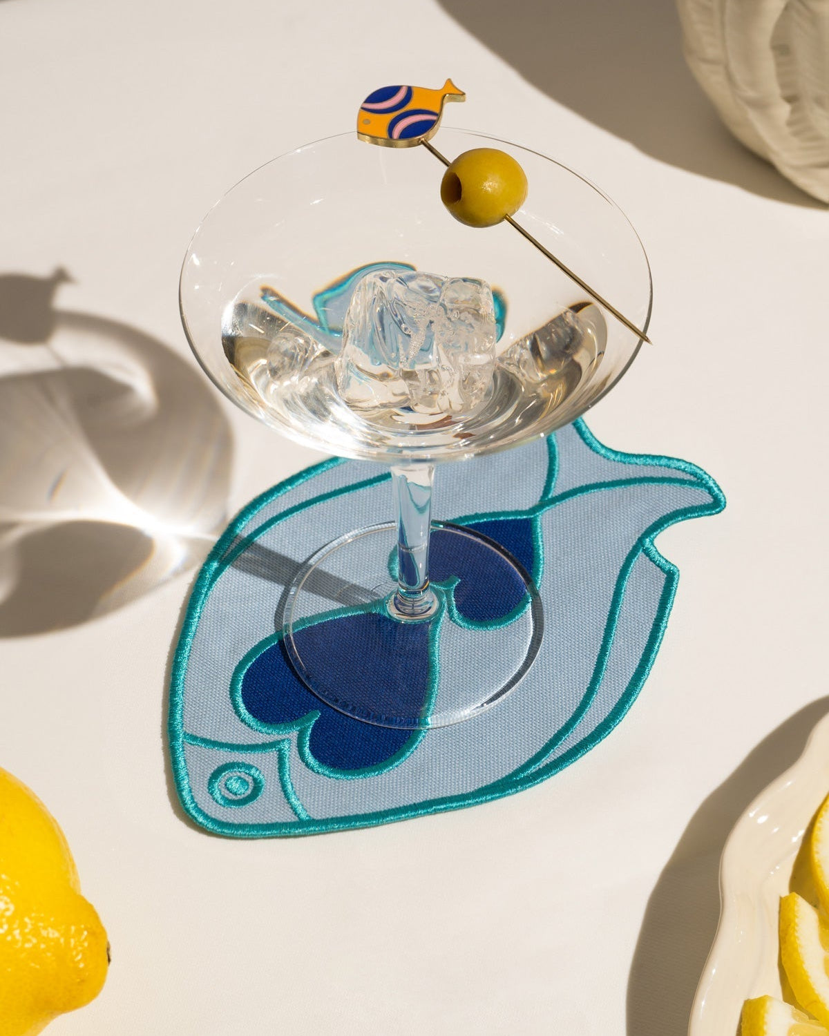 A martini with an olive rests on an Octaevo Fish Splash Coaster, beside a lemon and its fish-shaped shadow.