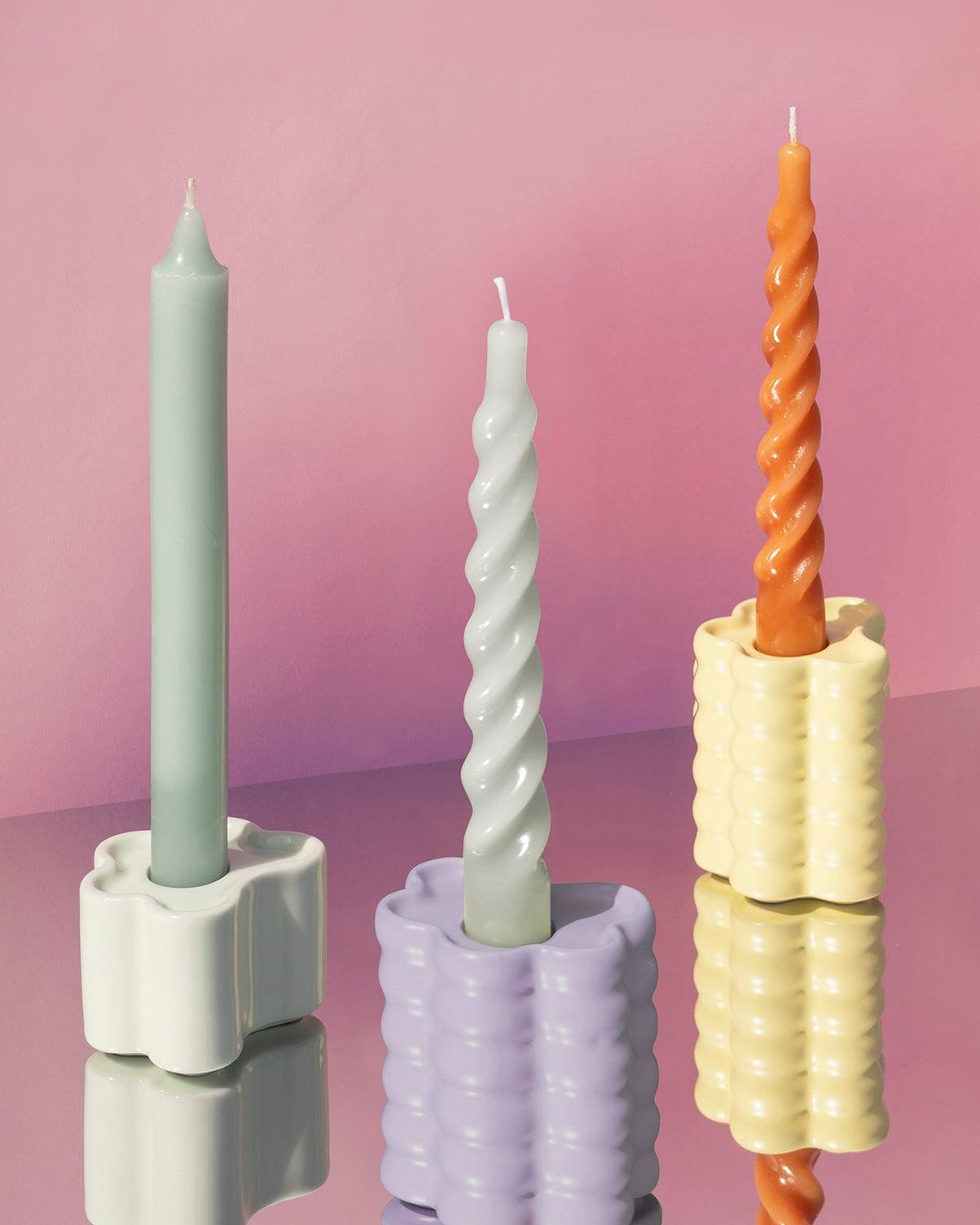 Flora Candle Holders by Octaevo with pastel candles are shown on a reflective surface against a pink background.
