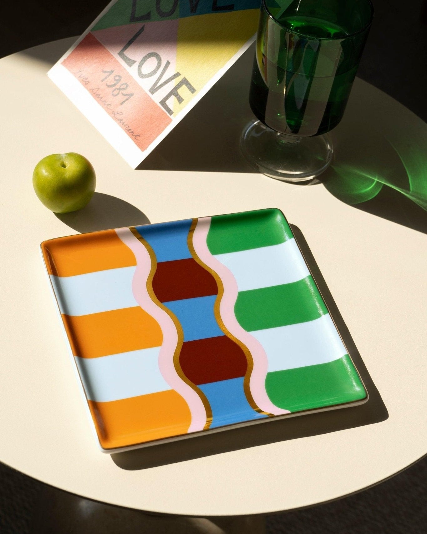 The OCTAEVO Giza Ceramic Tray with a green apple and LOVE card on a white table in sunlight.