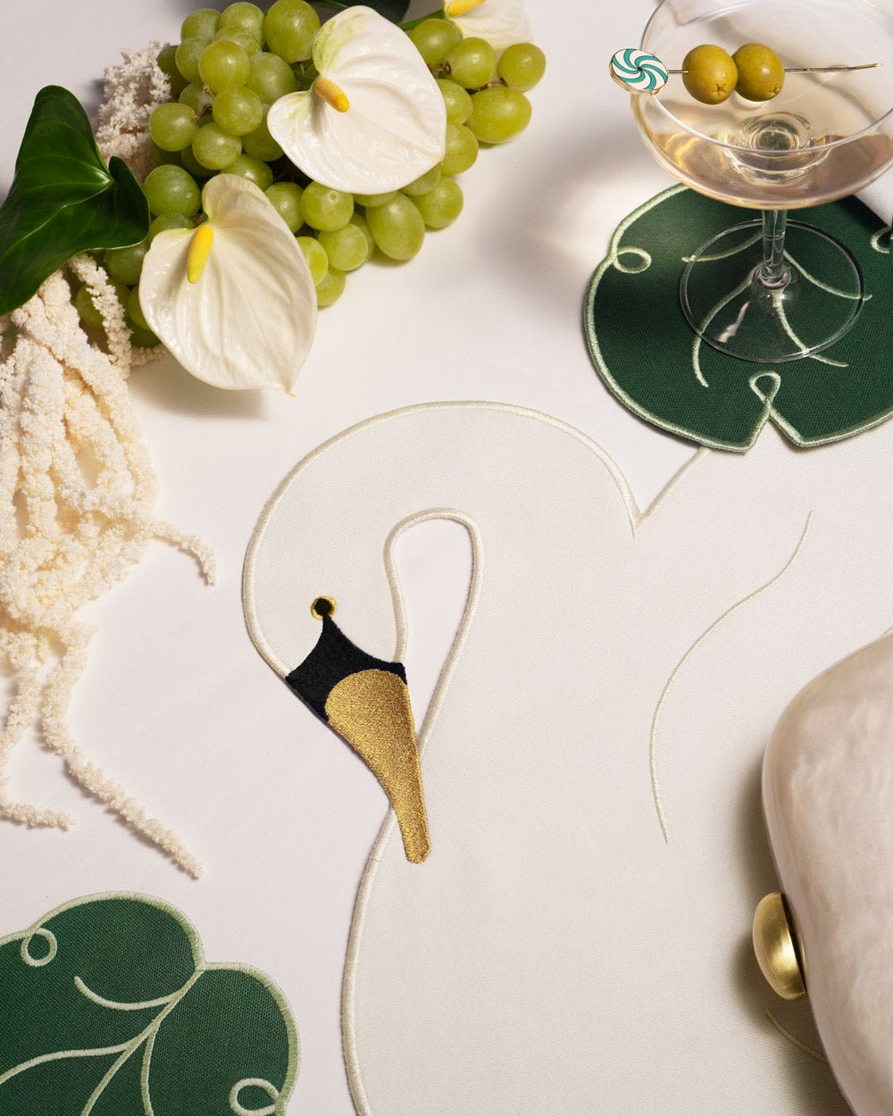 A white table setting with Octaevo Lily Leaf Coasters, a swan motif, grapes, white flowers, and a martini with olives.