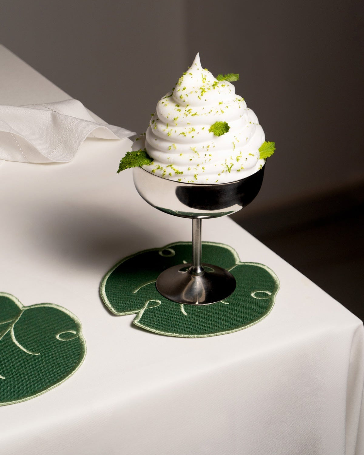 A metal cup with whipped dessert sits beside Octaevo’s Lily Leaf Coasters (Set of 4) on a white tablecloth.