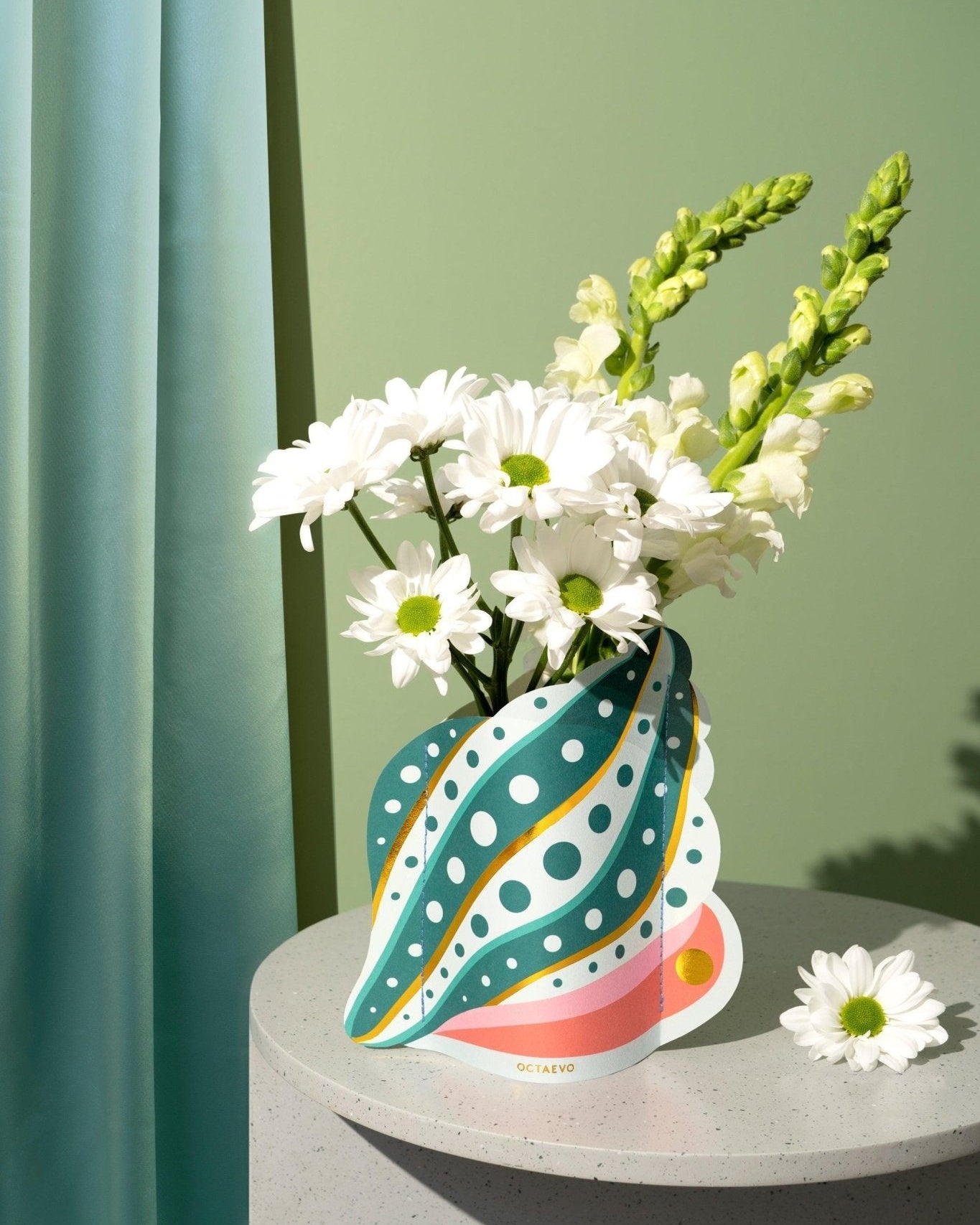 White daisies and snapdragons in the Octaevo Shell Mini Paper Vase, displayed on a gray table with a green background.