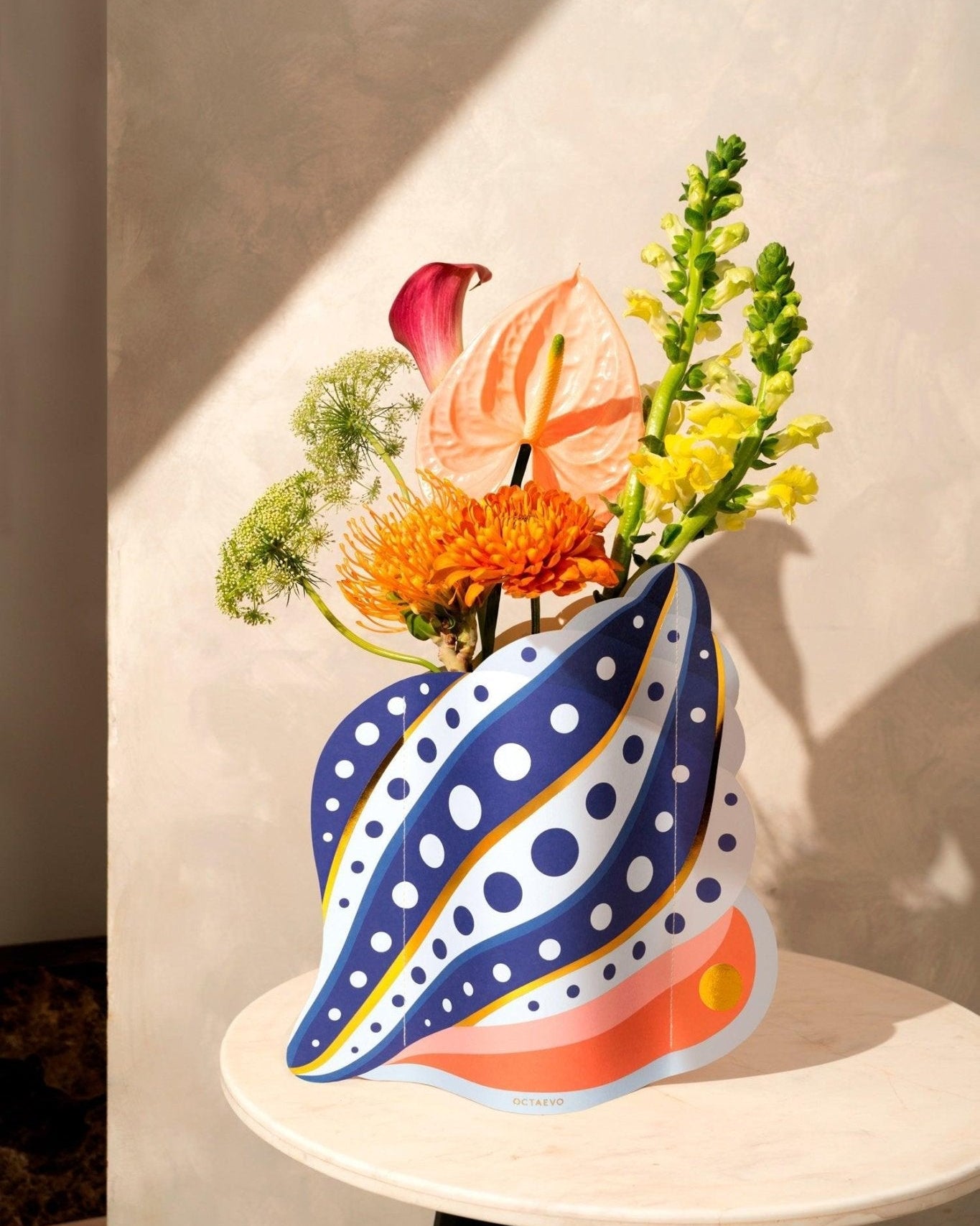 Vibrant flowers in Octaevos Shell Paper Vase create a Mediterranean centerpiece on a round table with a neutral backdrop.