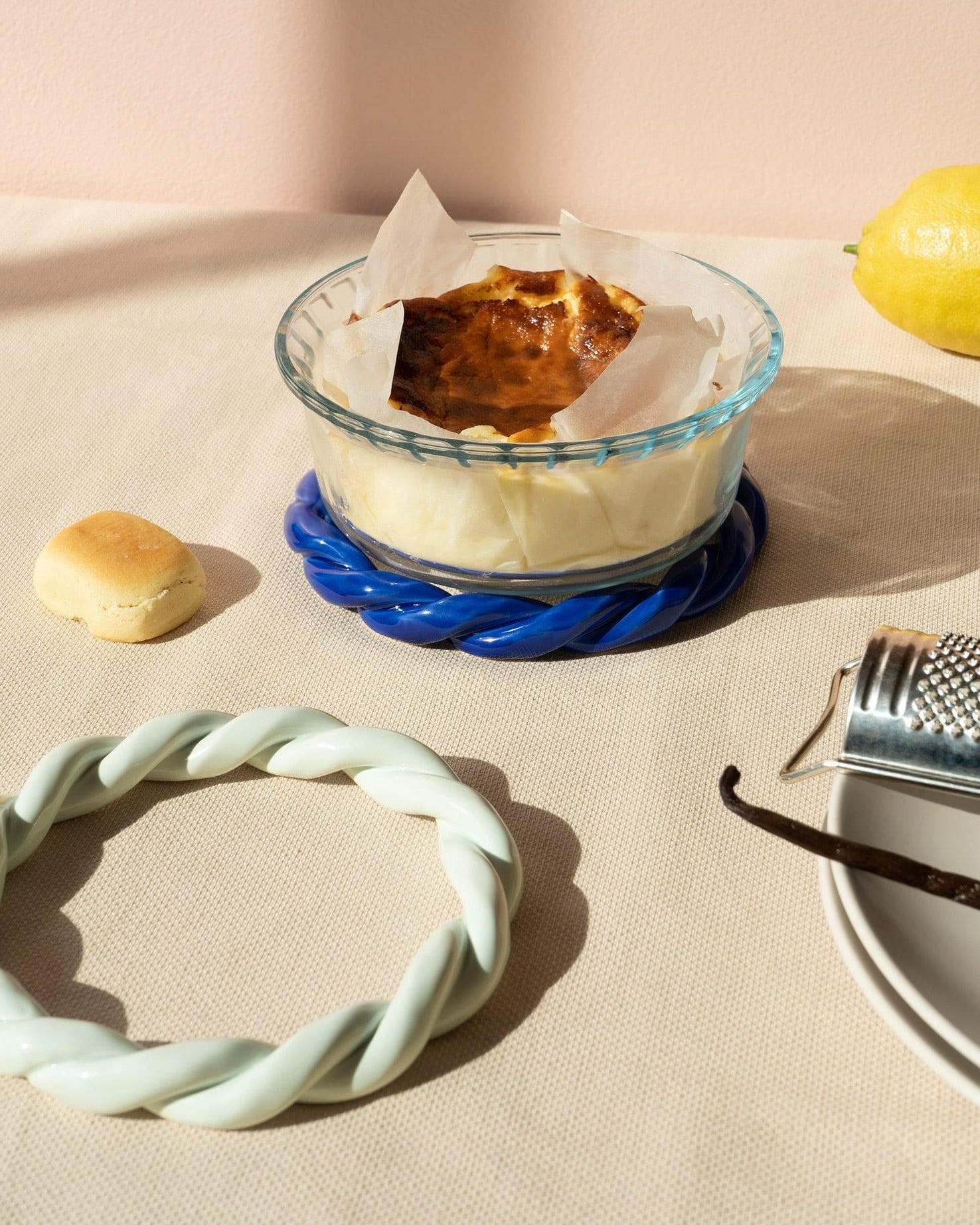A baked custard dessert in a glass dish on a light mint trivet, with a lemon, a roll, and a vanilla bean nearby.