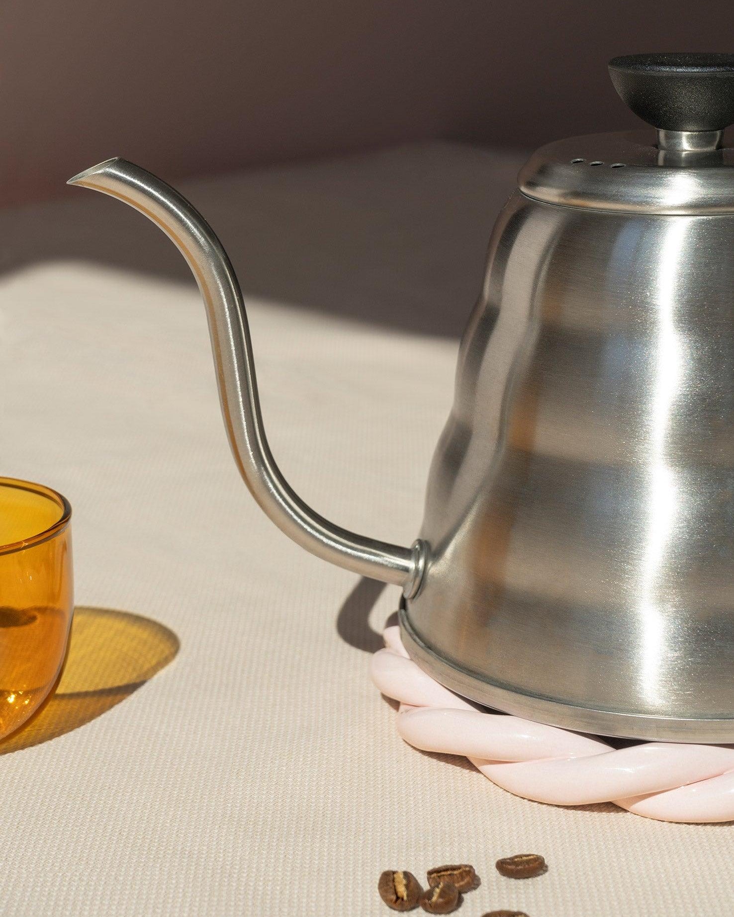 A pink OCTAEVO Sicilia Ceramic Trivet holds a gooseneck kettle near a yellow glass and coffee beans on beige.