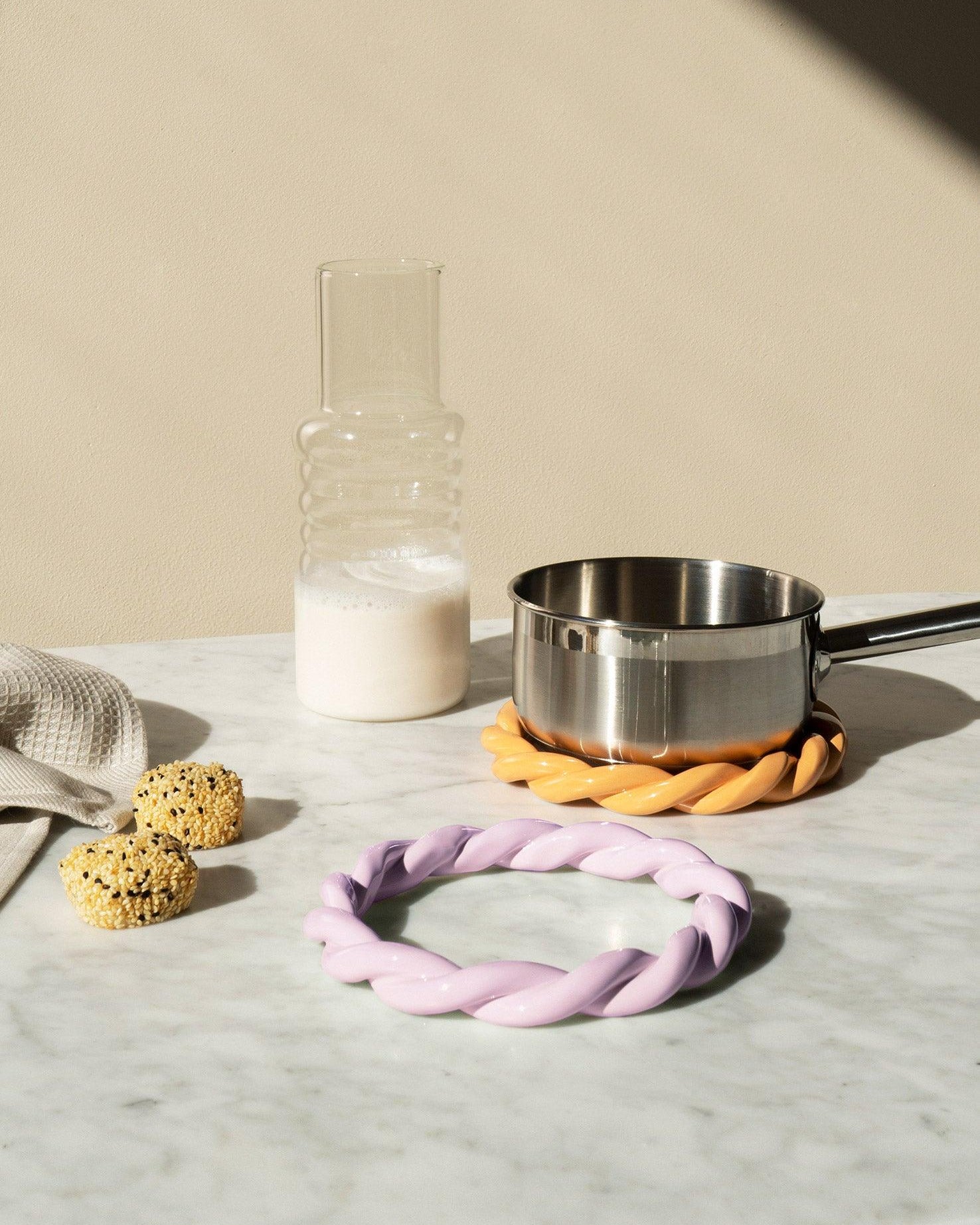 A glass carafe of milk, a saucepan on orange OCTAEVOs Sicilia Ceramic Trivet, and two snacks on a marble table.