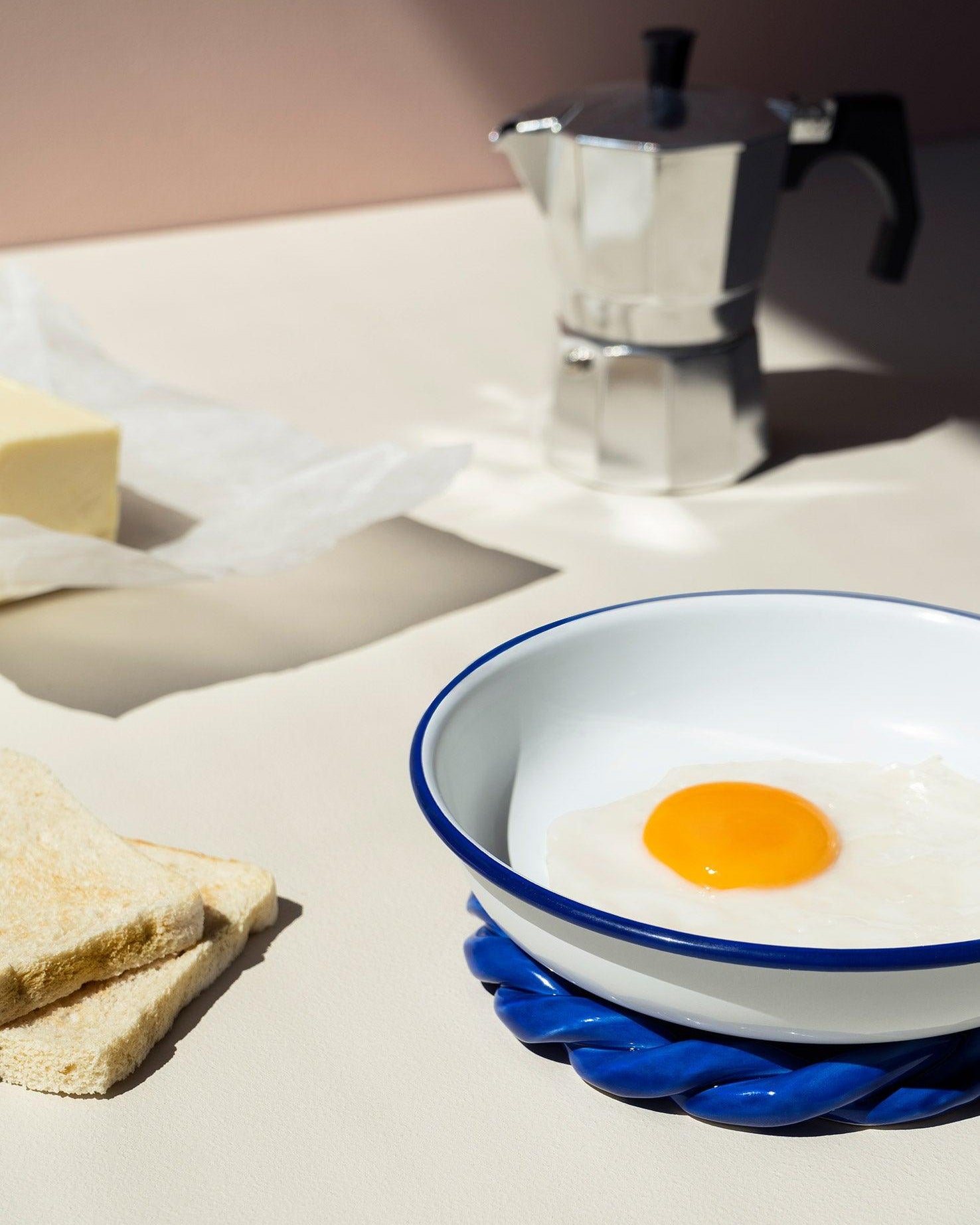 A bowl, toast, egg, butter, and a stovetop espresso maker sit on a blue OCTAEVO Sicilia Ceramic Trivet.