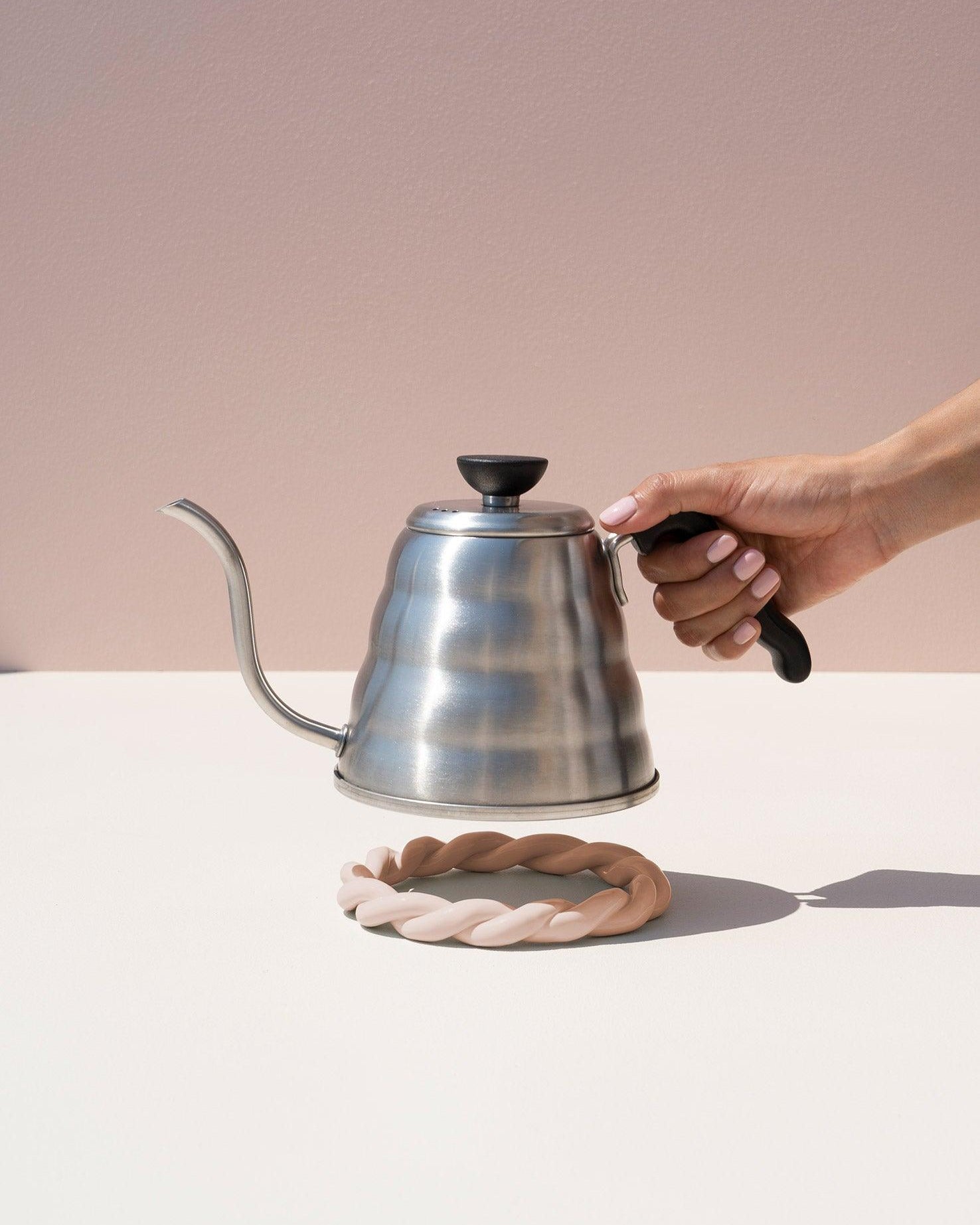 A hand holds a metal kettle above an OCTAEVO Sicilia Ceramic Trivet on a light surface with a pink background.