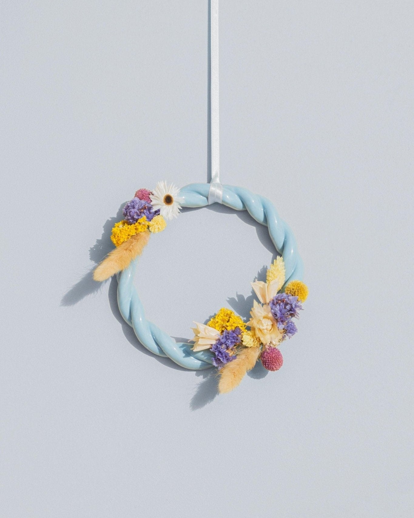 The light blue OCTAEVO Sicilia Ceramic Trivet is displayed against a light gray wall, hanging by a white ribbon.