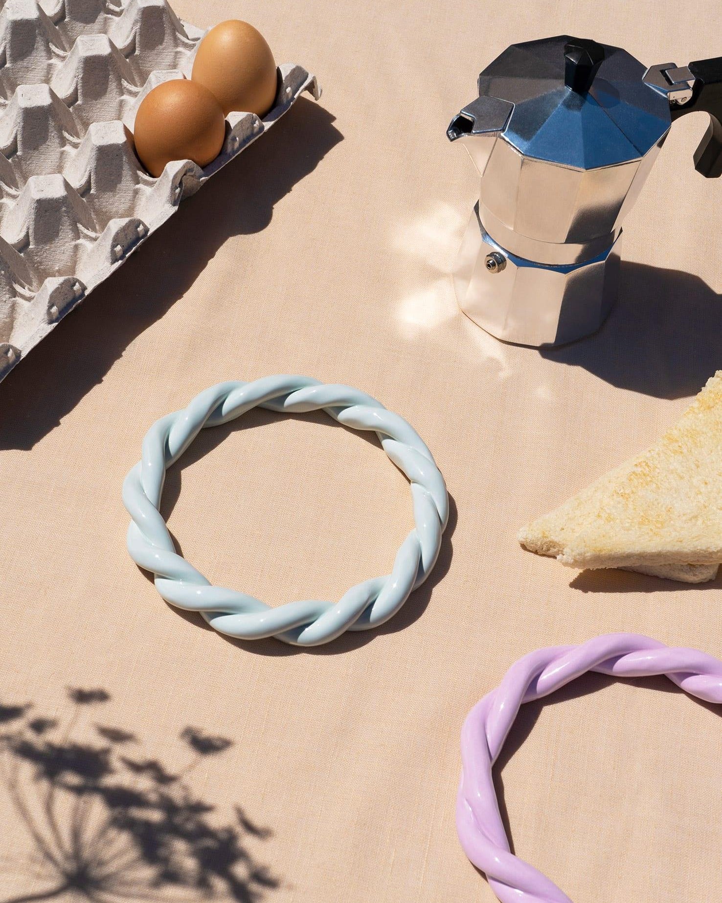 A moka pot, eggs, cheese wedge, and a light blue OCTAEVO Sicilia Ceramic Trivet on a beige tablecloth.