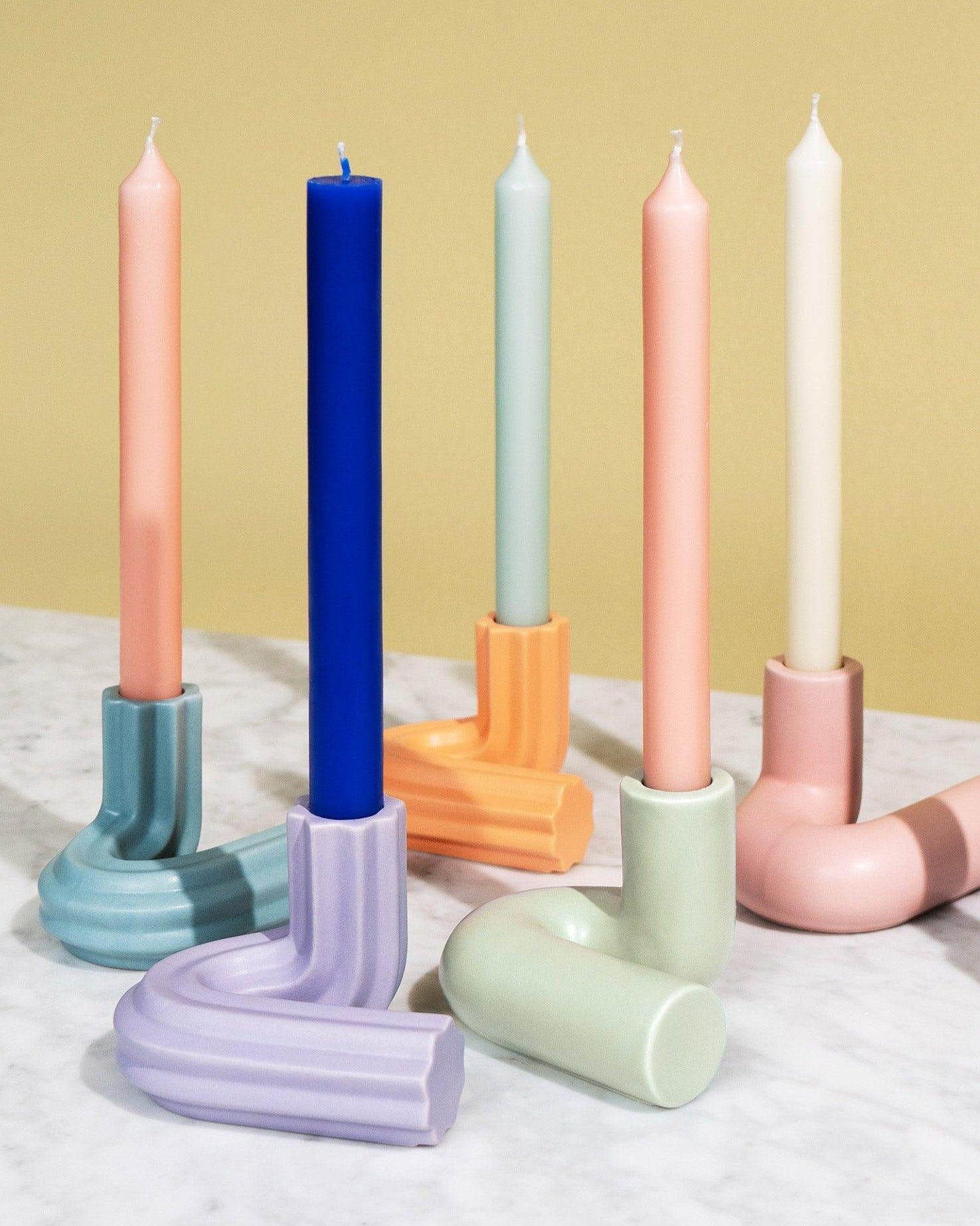 Five OCTAEVO Templo Candle Holders with colorful candles are arranged on a marble surface against a beige backdrop.