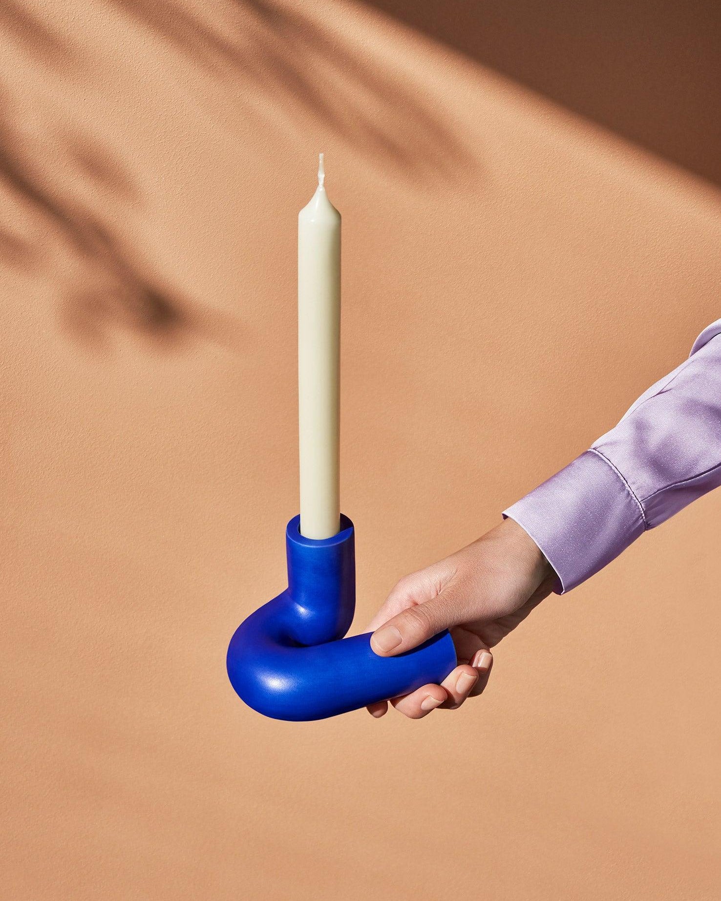 A hand holds the OCTAEVO Templo Candle Holder in blue with a white candle against a peach background.