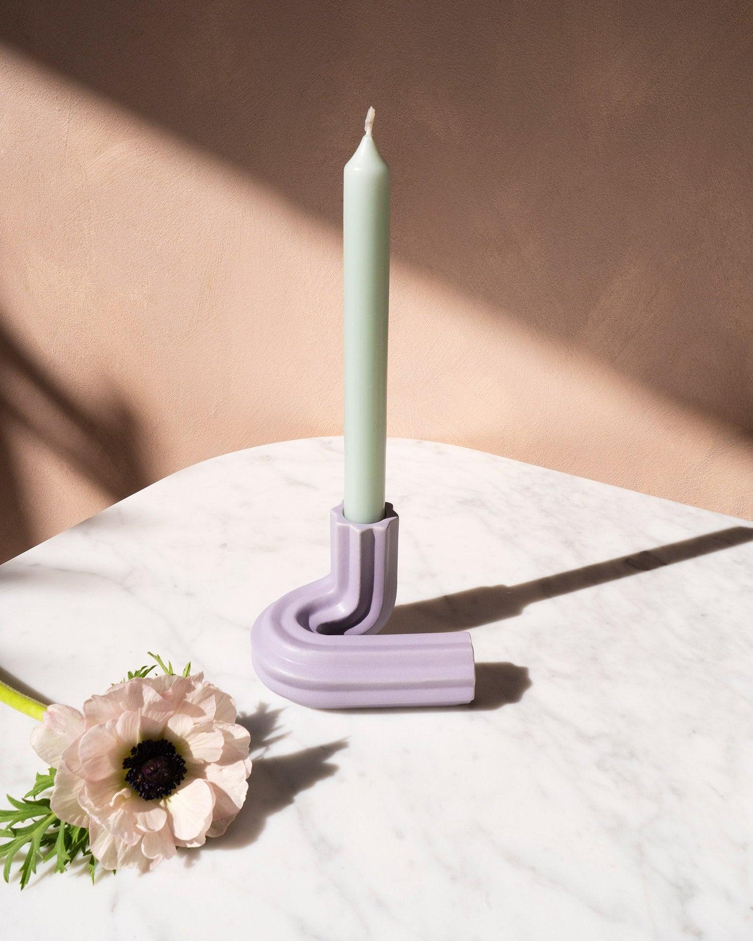 A pastel candle in OCTAEVO’s purple Templo Candle Holder, on marble, with a pale flower beside it.