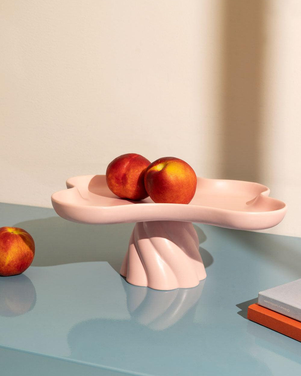 Three peaches on a pink, sculptural pedestal dish, with another peach and books on a glossy blue tabletop.