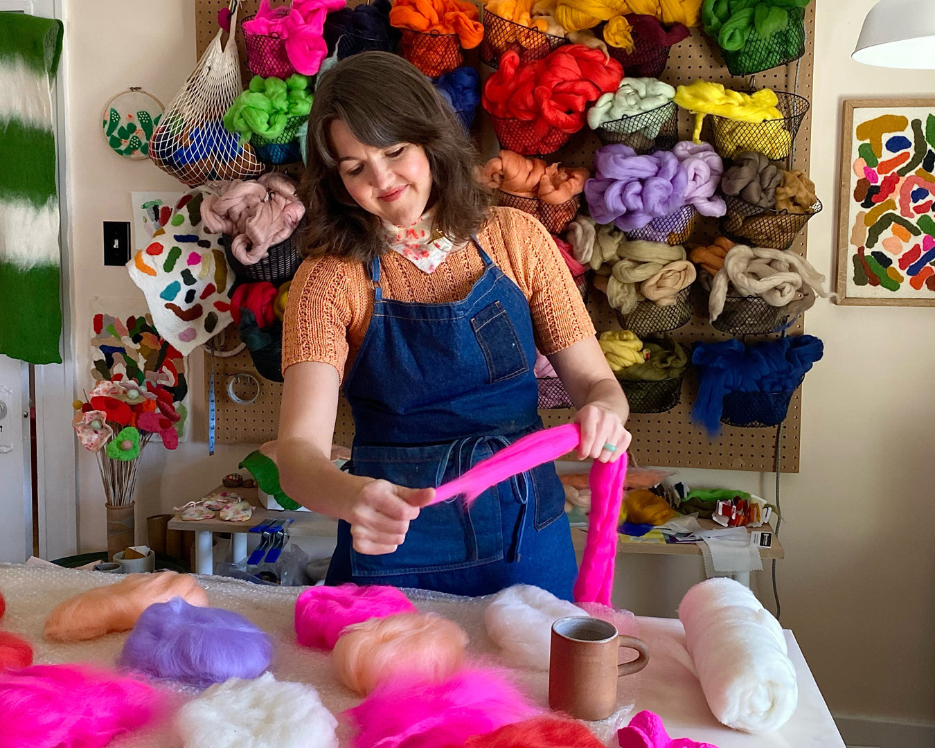 Artisan stretching vibrant pink wool during water felting workshop, surrounded by a rainbow of wool supplies. 