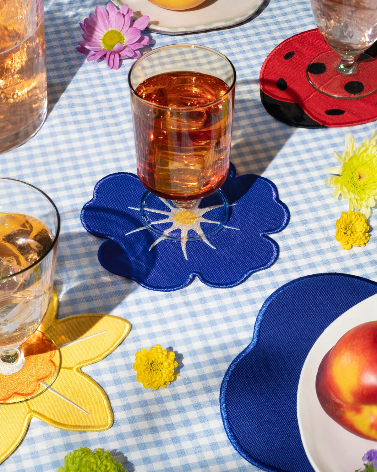 A glass rests on an Octaevo Morning Glory Coaster, atop a blue gingham tablecloth with flowers and peaches.