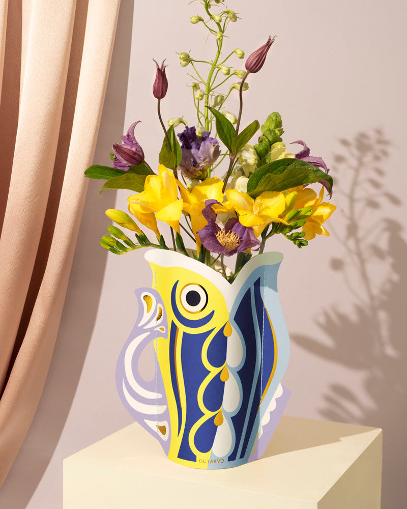 The Fish Mini Paper Vase by OCTAEVO with an abstract bird design holds flowers beside a light pink curtain.