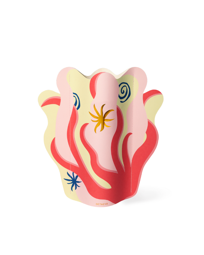 The Octaevo Amalfi Paper Vase features vibrant patterns and holds white and pink flowers on a beige table.