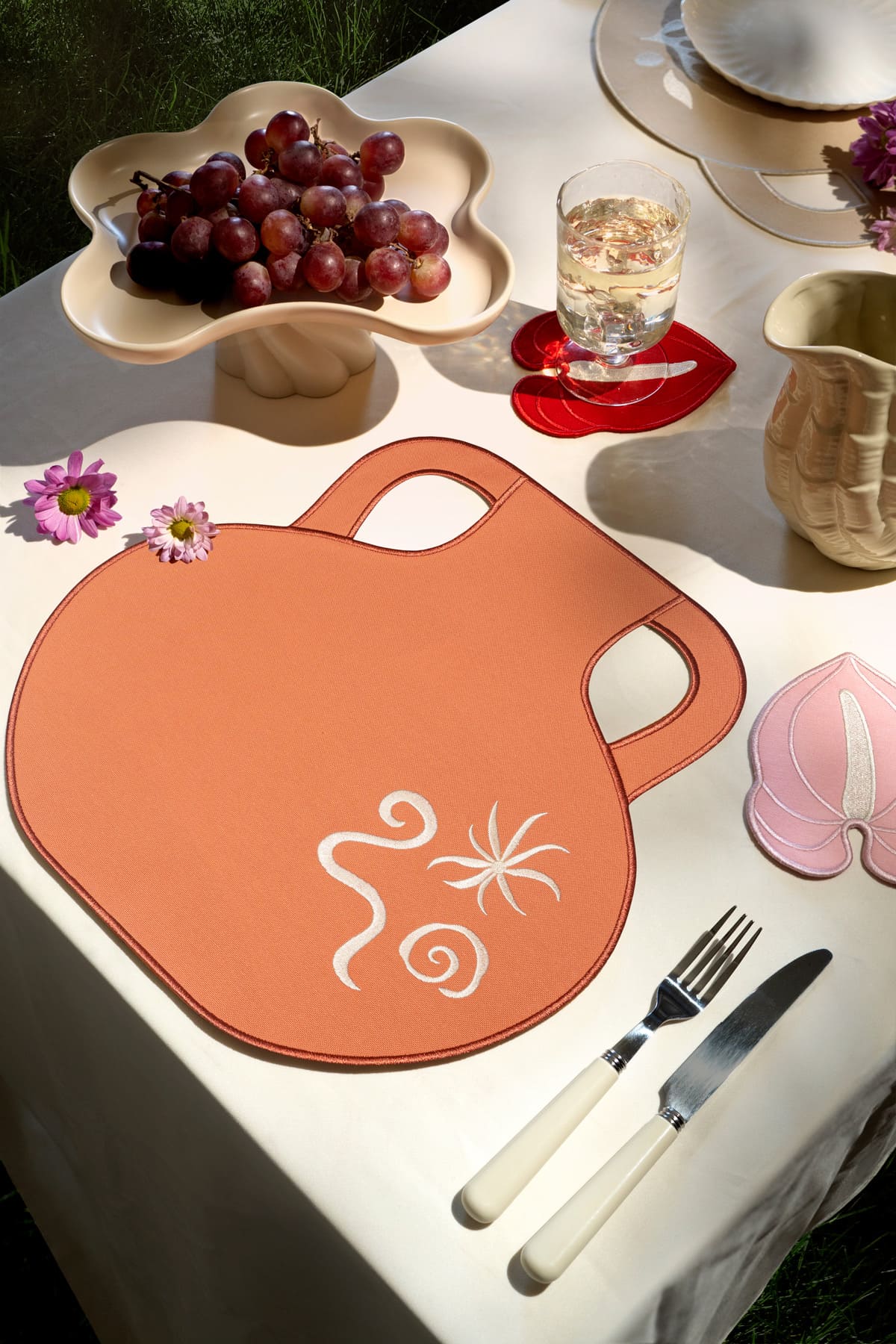 An outdoor table with grapes, a drink, flowers, and Octaevo’s Terracotta Amphora Placemats (Set of 2).