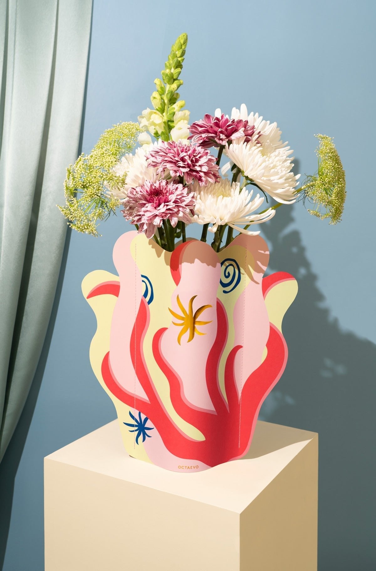 The Amalfi Paper Vase by Octaevo features assorted blooms on a pedestal, set against a blue Mediterranean backdrop.