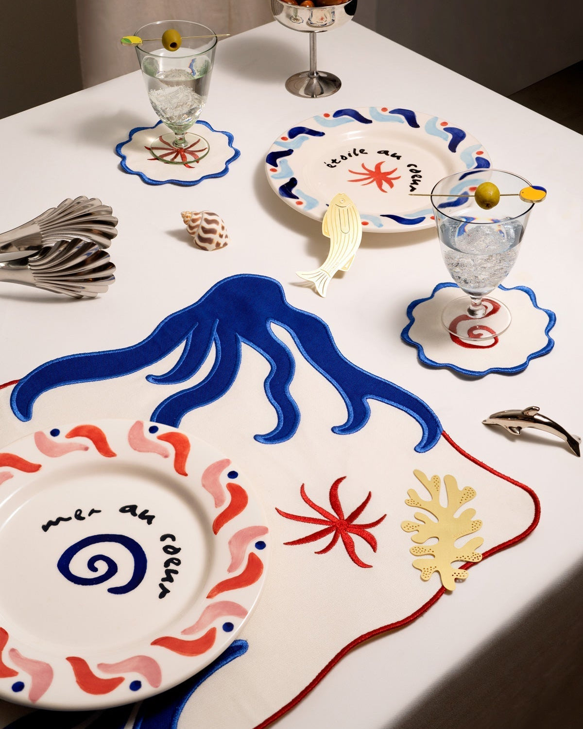 A table set with ocean plates, OCTAEVO Amalfi Placemats (Set of 2), olive glasses, and seashell decor.