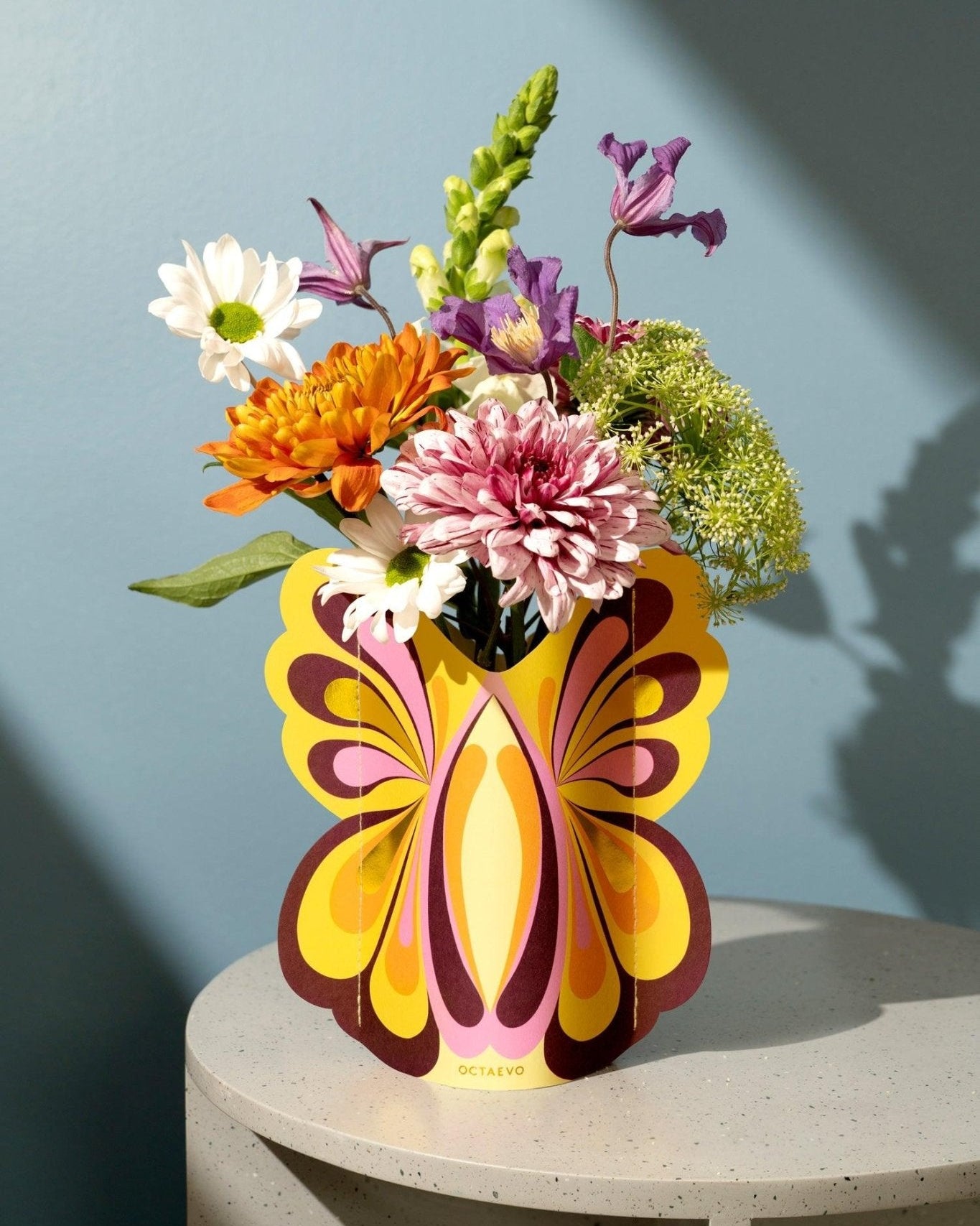 A vibrant bouquet in Octaevos Butterfly Mini Paper Vase rests on a speckled table against a blue wall.