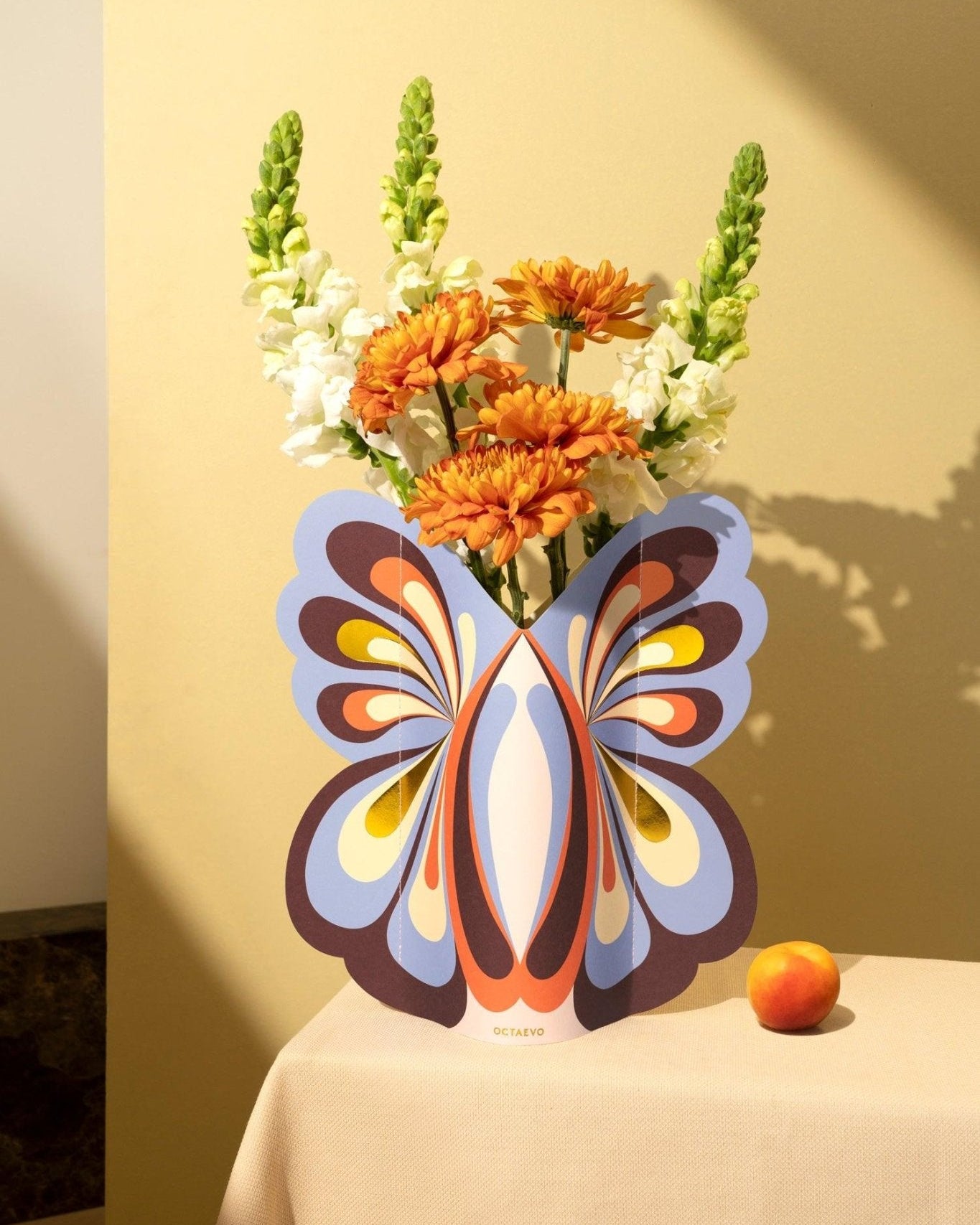 The Octaevo Butterfly Paper Vase holds flowers on a beige table, with a peach beside it.