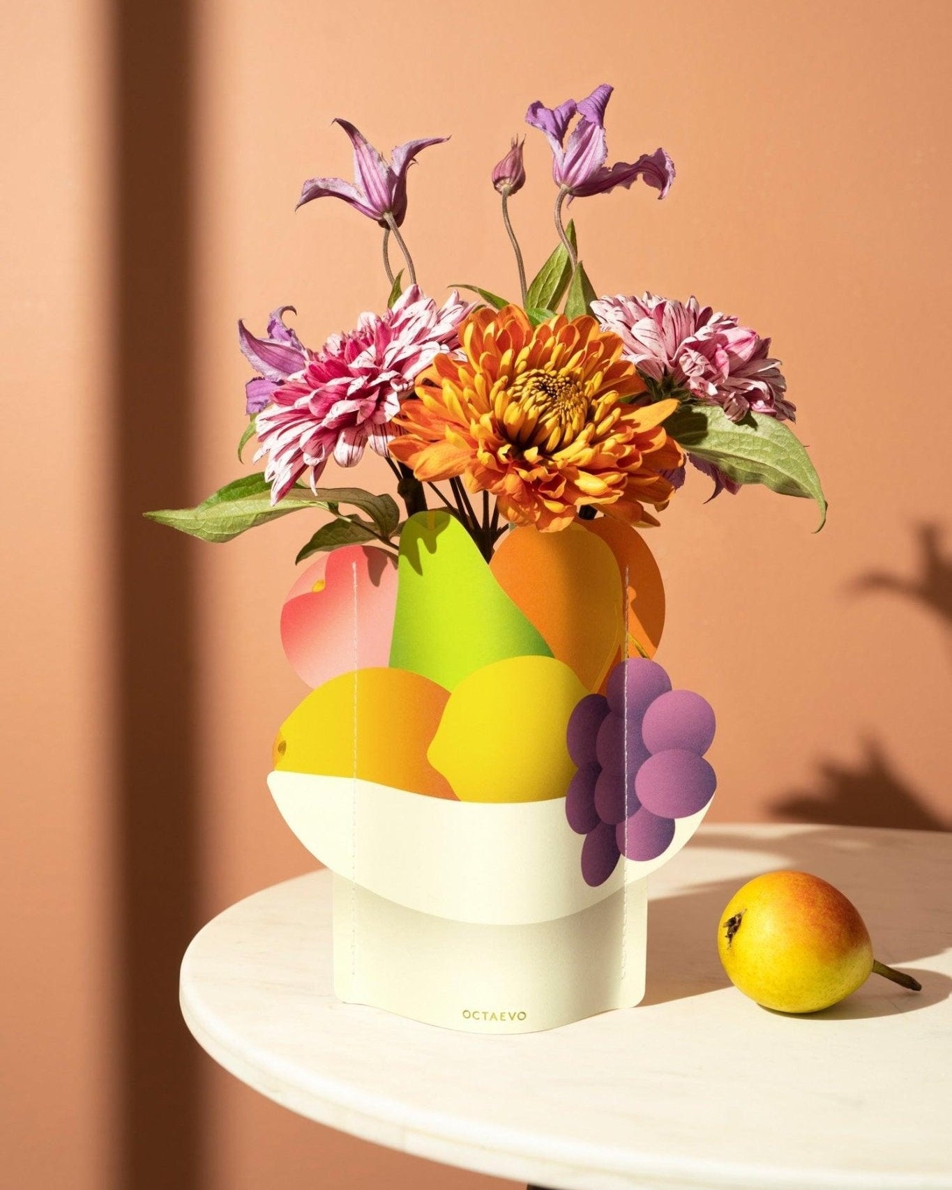 The Octaevo Fruit Bowl Mini Paper Vase with vibrant flowers sits on a white table against a peach-colored wall.