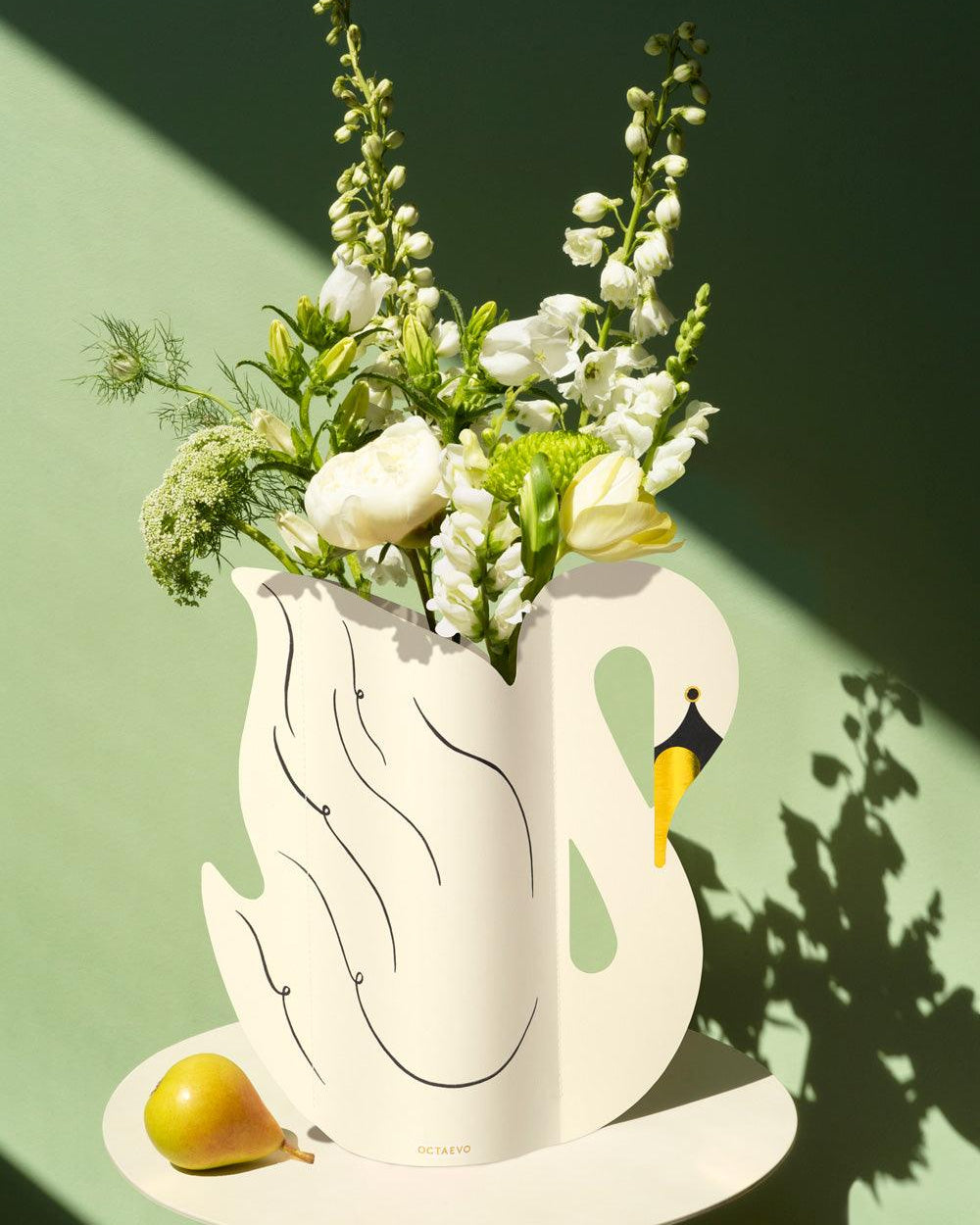 The Octaevo Swan Paper Vase with white flowers is displayed beside a yellow pear on a round table by a green wall.