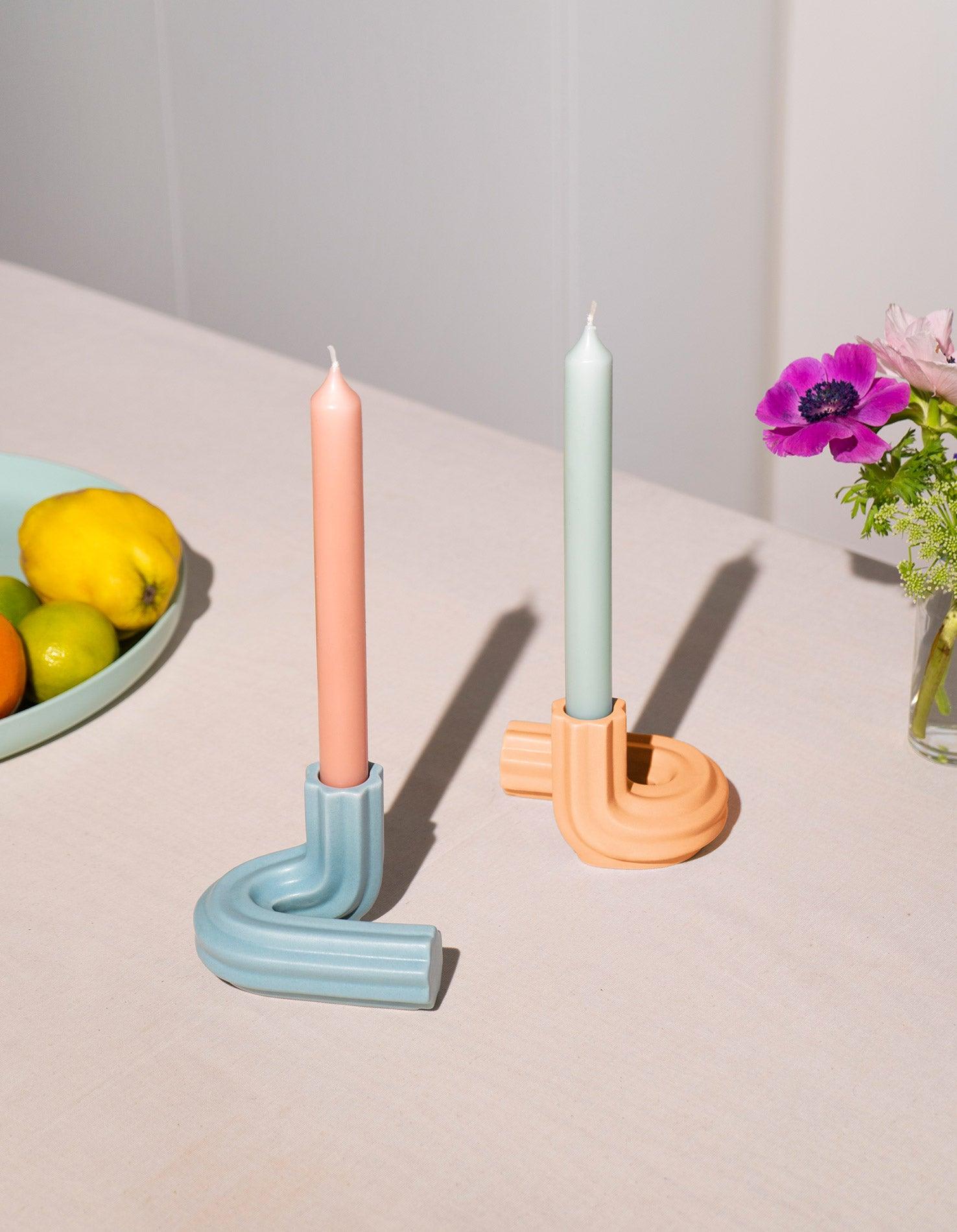 Two pastel candles in orange OCTAEVO Templo Candle Holders on a table, with fruit and flowers in the background.