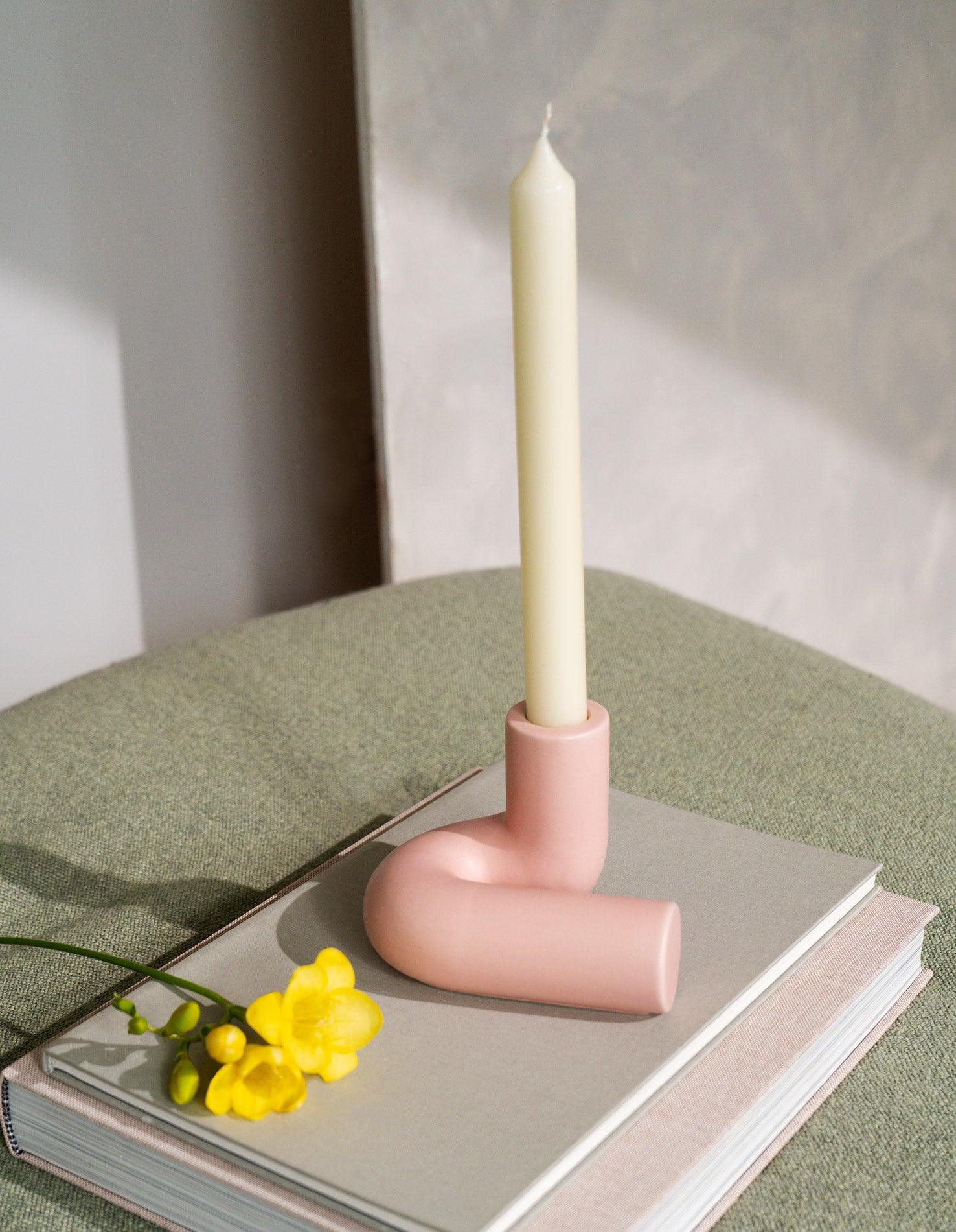 The OCTAEVO Templo Candle Holder in pink holds a white candle on a book beside a yellow flower, on light green surface.